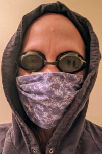 Person in a hoodie with swim goggles and a cloth over their mouth.